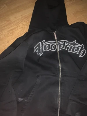 Svart zip hoodie från Hoodrich - Säljer en svart hoodie med dragkedja från Hoodrich. Stor logga i vitt och grått broderat över bröstet. Klassisk huva och två fickor framtill. Perfekt för en streetwear-look och riktigt skön att bära.
