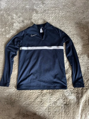 Mörkblå Nike träningströja med dragkedja - Mörkblå långärmad träningströja från Nike med vit rand över bröstet och liten dragkedja vid halsen. Samma passform som en S trotts barnstorlek. 
