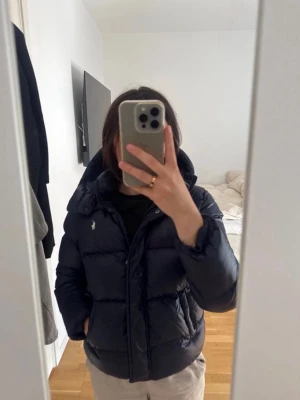 Mörkblå pufferjacka från Polo Ralph Lauren - Säljer en mörkblå pufferjacka från Polo Ralph Lauren med huva och dragkedja. Jackan har två sidofickor, hög krage och den klassiska lilla loggan broderad på bröstet. Perfekt för kalla vinterdagar och riktigt skön att bära.