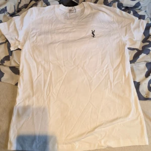 Vit t-shirt från Yves Saint Laurent - Klassisk vit t-shirt från Yves Saint Laurent med diskret YSL-logga broderad på bröstet. T-shirten har rund hals och korta ärmar, perfekt för en clean och stilren look. Tillverkad i mjuk bomull för maximal komfort.
