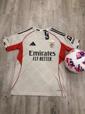 Benfica matchtröja Adidas beige/röd - Snygg Benfica matchtröja från Adidas i beige med röda och svarta detaljer. Tröjan har klubbmärke, sponsortryck och svarta ränder på axlarna. Tillverkad i lätt och ventilerande funktionsmaterial, perfekt för fotbollsträning eller match.