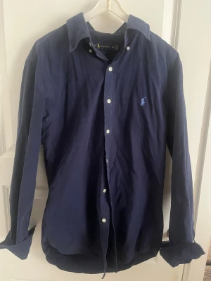 Mörkblå skjorta från Ralph Lauren - Klassisk mörkblå skjorta från Ralph Lauren med vit knappstängning och broderad logga på bröstet. Skjortan har lång ärm, normal passform och är tillverkad i mjuk bomull. Perfekt för en stilren och avslappnad look.
