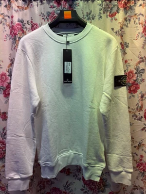Vit sweatshirt från Stone Island - Säljer en clean vit sweatshirt från Stone Island med klassisk rund hals och ribbade muddar. Tröjan har den ikoniska svarta logotypen på vänster ärm och är tillverkad i mjukt bomullsmaterial. Perfekt för dig som gillar stilrena och trendiga plagg.