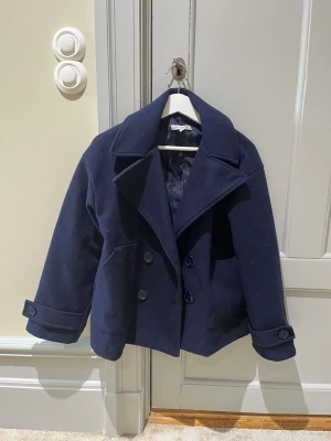 Marinblå kort kappa från DesignbySi - Stilren marinblå kappa dubbelknäppning och stora slag. Jackan har två framfickor och dekorativa knappar vid ärmsluten. Perfekt för dig som vill ha en klassisk look med modern touch.