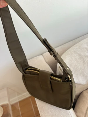 Olivgrön axelväska - Snygg olivgrön axelväska med justerbart axelband och guldfärgade metalldetaljer undertill. Väskan har dragkedja upptill och en stilren, rektangulär form som passar till många outfits.