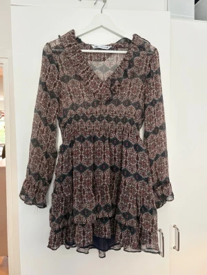 Blommig volangklänning från Zara - Supersöt klänning från Zara med volangdetaljer och lång ärm. Klänningen har ett mönster i blått, vinrött och beige, smock i midjan och volanger upptill och nertill. 