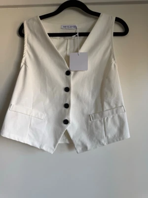 Vit väst med knappar från New Collection - Stilren vit väst från New Collection med djup V-ringning och fyra svarta knappar framtill. Västen har två dekorativa fickor och är tillverkad i ett slätt tyg som ger en clean look. Perfekt för att lyfta din outfit med en modern touch.