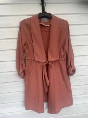 Rostrosa trenchcoat med knytskärp - Trendig rostrosa trenchcoat med avslappnad passform och knytskärp i midjan. Jackan har bred krage, uppvikta ärmar med knappdetalj och två fickor framtill. Perfekt för lager på lager och ger en cool vibe till din outfit.