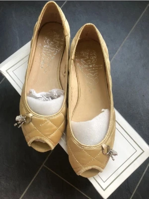Beige quiltade ballerinaskor med charm 37  - Snygga beige ballerinaskor från Doretha med quiltat mönster och en söt charm framtill. Skorna har en öppen tå och är perfekta för en stilren look. Färgen är light camel