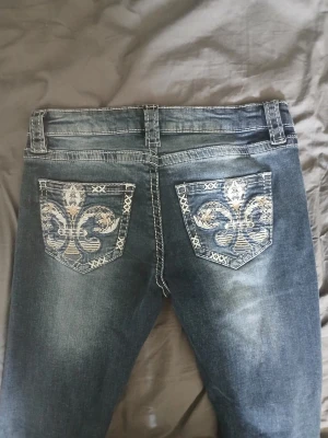 Mörkblå jeans med broderade bakfickor - Snygga mörkblå jeans från Grace in LA med slitningar och kontrastsömmar. Bakfickorna har stora broderade mönster i vitt och beige som ger en unik look. Klassisk femficksmodell med normal midja och smal passform. Perfekta för dig som vill sticka ut.