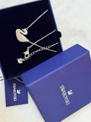 Swarovski halsband med svan och kristaller - Supersnyggt halsband från Swarovski med en silverfärgad kedja och ett svanhänge täckt av gnistrande vita kristaller. Hängsmycket har en elegant form och detaljerade stenar som ger extra bling. Levereras i originalask med märkets logga.