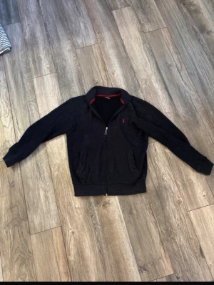 Svart ziptröja från Ralph Lauren - Snygg svart ziptröja från Ralph Lauren med röd broderad logga på bröstet och röda detaljer i kragen. Tröjan har två fickor framtill, ribbade muddar och hög krage. Perfekt för en chill och stilren look. Det står storlek M men den är mer XS