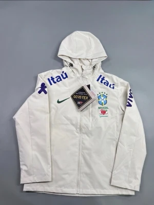 Nike Brasil vindjacka med huva - Snygg beige vindjacka från Nike med Brasil-logga och sponsormärken i blått och grönt. Jackan har huva, dragkedja framtill och är tillverkad i GORE-TEX-material som står emot vind och regn. Perfekt för dig som gillar sportig stil och vill sticka ut.