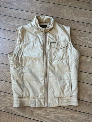 Wrangler väst - Stilren beige dunväst med dragkedja framtill och en praktisk ficka med knappstängning på ena sidan. Västen har quiltad design och är perfekt för lager-på-lager-look. 