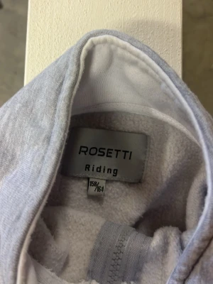Ljusgrå hoodie från Rosetti Riding - Ljusgrå hoodie från Rosetti Riding med dragkedja framtill och två stora fickor. Mjuk insida i fleece och tryckt logga på bröstet. Perfekt för chill dagar eller när du vill ha något bekvämt. Klassisk huva och enkel, stilren design.