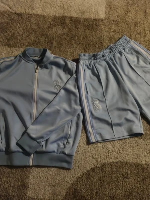 Ljusblått loungewear set från Risky - Snyggt ljusblått loungewear set från Risky med matchande zip-jacka och shorts. Setet har vita och blå ränder längs sidorna och broderad logga på både jacka och shorts. Tillverkat i mjukt, glansigt material med elastisk midja på shortsen för extra komfort. Storleken Small herr, passar väldigt bra och ett allmänt bra val för ett sommar set denna fina sommaren. Fast pris!