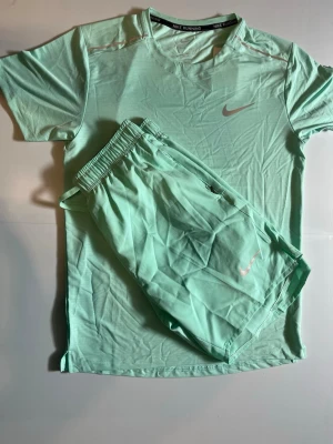 Mintgrönt träningsset från Nike - Snyggt mintgrönt träningsset från Nike med t-shirt och shorts. Båda plaggen har Nike-logga och är gjorda i ett lätt, snabbtorkande material som passar perfekt för löpning eller gym. T-shirten har rund hals och shortsen har resår i midjan.