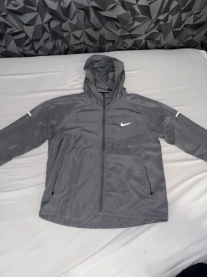 Nike Miller jacka  - Snygg grå vindjacka från Nike med huva och dragkedja framtill. Jackan har två fickor med dragkedja och reflekterande detaljer på ärmarna. Perfekt för löpning eller promenader. Tillverkad i lätt och slitstarkt syntetmaterial. Jackan är aldrig använd och endast testad 