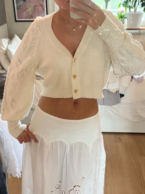Vit cropped kofta med puffärm - Supersöt vit cropped kofta med puffiga ärmar och fina hålstickade detaljer. Koftan har v-ringning, ribbade muddar och tre guldknappar framtill. Perfekt att styla över klänning eller linne för en trendig look.