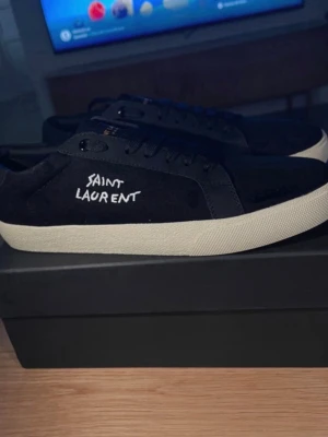 Svarta Saint Laurent sneakers - Svarta sneakers från Saint Laurent med vit broderad logga på sidan och svart läderdetalj vid hälen. Skorna har en vit, tjock sula och är tillverkade i en mix av mocka och läder. Klassisk låg modell med svarta skosnören och stilren design.
