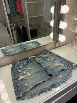 Blå jeanskjol med slitningar - Säljer en kort jeanskjol från One Teaspoon i ljusblå denim med slitna detaljer och fransig kant. Kjolen har klassisk femficksmodell, knappgylf och råa kanter för en edgy look. Perfekt för dig som gillar streetstyle och avslappnad vibe.