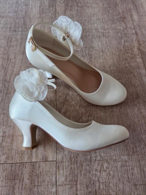 Satinskor i pumpsmodell med blomdeko och rem 38 - Satinskor i pumpsmodell med avtagbar blomdekoration och med rund tå medelhög klack. Skorna har en justerbar ankelrem med guldfärgat spänne Material lyxig satin. Klackhöjd ca 6 cm