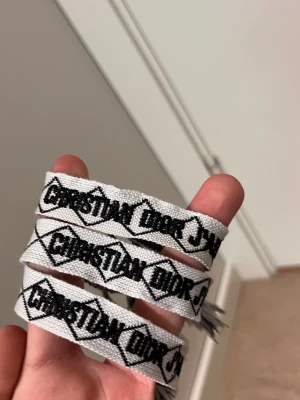 Vitt tygarmband Christian Dior - Säljer ett vitt tygarmband från Christian Dior med svart broderad logga. Armbandet har en klassisk, vävd design och avslutas med fransar. Perfekt accessoar för att lyfta din outfit med en exklusiv touch.