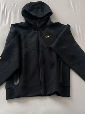Svarta NOCTA x Nike dress  - Säljer ett par svarta mjukisbyxor från Nike och NOCTA-kollektionen. Byxorna har gul logga, dragsko i midjan och en unik ficka med dragkedja på sidan. Broderade detaljer i gult på benen och relaxed passform. Perfekta för chill eller streetwear.