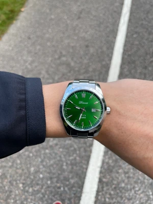 Grön klocka med stållänk från Doventi  - Snygg klocka från Doventi med grön urtavla och datumvisning. Klockan har en klassisk rund form och en stilren länk i silverfärgat stål. Perfekt accessoar för dig som vill sticka ut med en färgglad detalj på handleden.