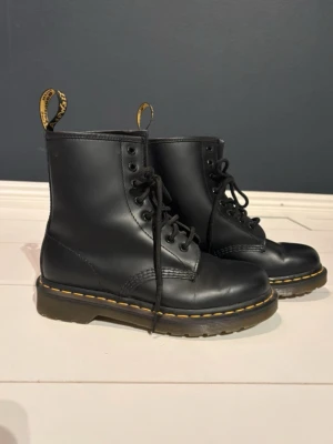 Svarta Dr. Martens kängor i läder - Klassiska svarta Dr. Martens kängor i slätt läder med snörning och den ikoniska gula sömmen runt sulan. Grov sula och dragflik bak med logga. Perfekta för dig som vill ha en tidlös och edgy stil.