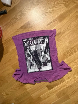 Lila t-shirt med New York-tryck - Cool lila t-shirt med stort grafiskt tryck av New York på framsidan. T-shirten har klassisk passform och korta ärmar. Perfekt för dig som gillar streetstyle och vill sticka ut med en unik färg.