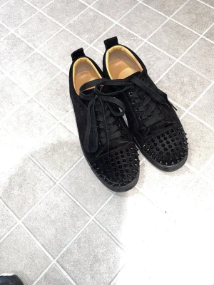 Svarta sneakers med nitar från Louboutin - Säljer ett par svarta sneakers från Christian Louboutin i mocka med svarta nitar på tån och klassisk röd sula med logga. Skorna har svarta skosnören och låg profil. Perfekta för dig som vill sticka ut med en edgy look.