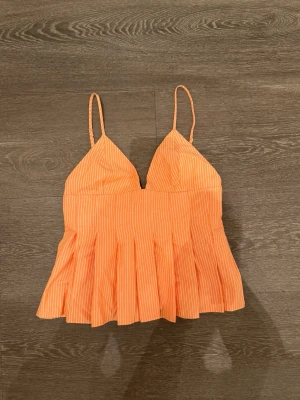 Randig orange sommartopp med tunna band - Superfin orange topp med vita smala ränder och tunna axelband. Toppen har v-ringning och volangliknande nederdel som ger en luftig känsla. Perfekt för varma dagar och enkel att matcha med jeans eller kjol. Liten i storleken
