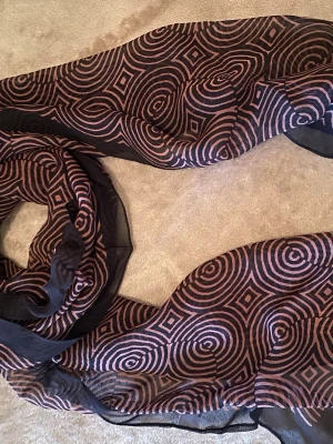 Brun sjal med rosa grafiskt mönster - Snygg och tunn svart sjal med rosa grafiskt cirkel- och fyrkantsmönster. Tillverkad i 100% polyester, vilket gör den lätt och luftig. Perfekt att slänga över axlarna för en cool touch till din outfit.