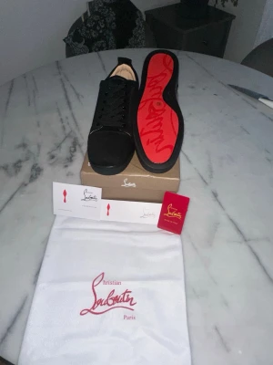 Svarta Christian Louboutin sneakers - Snygga svarta sneakers från Christian Louboutin med klassisk röd sula och diskret logga. Skorna har en låg profil, svarta skosnören och är tillverkade i mocka och läder. Perfekta för dig som vill ha en exklusiv och stilren look.