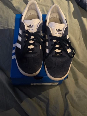 Adidas Gazelle mörkblå sneakers - Klassiska Adidas Gazelle sneakers i mörkblå mocka med vita detaljer och de ikoniska tre ränderna på sidan. Låg modell med svart snörning och vit sula. Perfekta för dig som gillar retrostil och streetwear.
