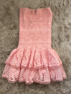 Rosa volangklänning från Designbyamor - Supersöt rosa kortklänning från Designbyamo med smockad överdel och två lager volanger nertill. Klänningen är ärmlös och har fina broderade detaljer i tyget som ger en romantisk känsla. Perfekt för sommarens alla tillfällen! Använd fåtal gånger
