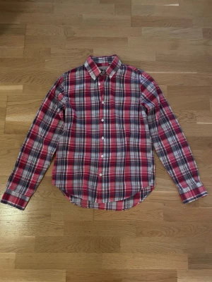 Rutig skjorta från Velour  - Klassisk röd, blå och vit rutig skjorta från Velour by Nostalgia i storlek XS. Skjortan har lång ärm, knappar framtill och en tidlös krage. Perfekt för dig som gillar en avslappnad men ändå stilsäker look.