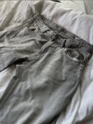 Ljusgrå jeans med rak passform - Säljer ett par ljusgrå jeans med klassisk femficksdesign och rak passform. Jeansen har bälteshällor, knappgylf och subtila slitningar vid fickan för en avslappnad look. Perfekta till en chill outfit.