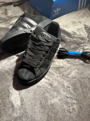 Svarta Adidas campus Originals sneakers - Snygga svarta Adidas Originals sneakers i mocka med klassiska tre ränder på sidan och Adidas-logga på plösen. Skorna har tjocka svarta skosnören och en låg siluett. Perfekta för dig som gillar stilrena och tidlösa sneakers.