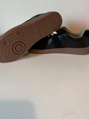 masion margiela - Stilrena svarta sneakers med ovandel i läder och mocka, vita snören och en klassisk brun gummisula. Skorna har lågt skaft och en tidlös design som passar till många outfits. Insidan är ljus och ger en snygg kontrast mot det svarta yttermaterialet.