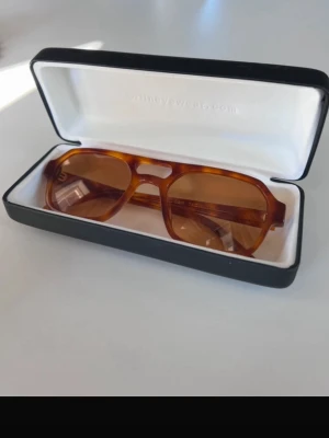 Bruna solglasögon från Corlin Eyewear - Snygga solglasögon från Corlin Eyewear med brunt, transparent båge i retroinspirerad fyrkantig form. Glasen är tonade i brunt och bågarna har en cool, bred design. Perfekta för att lyfta din stil under soliga dagar.