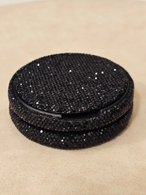 Svart glittrande Kelly White dosa - Snusdosa täckt med svarta glittrande stenar. Ytan är helt täckt av små svarta dekorstenar som ger en lyxig och iögonfallande look. Praktisk och snygg accessoar för dig som gillar glitter. Du lägger i dina påsar och har då en glittrande snygg snusdosa! Använd en gång. En eller 2 stenar har lossnat.