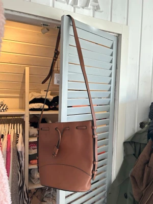 Brun bucket bag i läder - Snygg brun axelväska i bucket-modell med justerbar rem och dragskostängning. Väskan är gjord i slätt läder och har en innerficka för småsaker. Perfekt för dig som gillar stilrena och praktiska accessoarer.