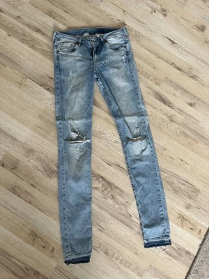 Ljusblå slitna skinny jeans - Säljer ett par ljusblå skinny jeans med slitningar och hål på knäna. Jeansen har klassisk femficksmodell, låg midja och råa kanter vid bensluten. Perfekta för en avslappnad och trendig look.