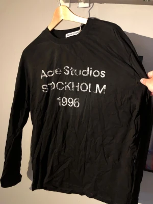 Svart långärmad t-shirt Acne Studios - Svart långärmad t-shirt från Acne Studios med vit text 'Acne Studios STOCKHOLM 1996' på bröstet. Klassisk rund halsringning och avslappnad passform. Perfekt för en stilren och trendig look.