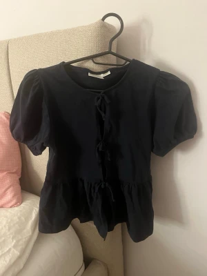 Svart blus med puffärmar och knyt - Säljer en blå blus med puffiga kortärmar och knyt framtill. Blusen har en peplum-liknande nederkant och är tillverkad i mjuk bomull. Perfekt för dig som gillar en söt och trendig look med volym och detaljer.