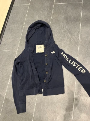 Mörkblå hoodie från Hollister Dam - Snygg mörkblå hoodie från Hollister med knäppning framtill och stor huva. Hollister-logga i vitt på ena ärmen och liten logga på bröstet. Tillverkad i mjukt bomullsmaterial, perfekt för en avslappnad och trendig look. Defekt (sista bilden): knappen längst ner saknas. Liten i storleken mer som (S/M)