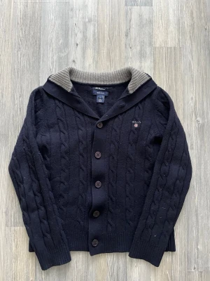 Mörkblå kabelstickad kofta från GANT - Säljer en mörkblå kabelstickad kofta från GANT i mjuk lammull. Koftan har knappar framtill, ribbade muddar och en grå ribbstickad insida på kragen. Broderad GANT-logga med sköld på bröstet. Perfekt för kyliga dagar!