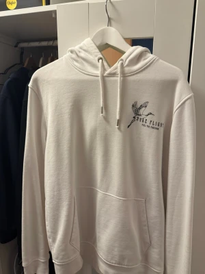 Vit hoodie med tryck - Säljer en vit hoodie med huva och känguruficka. På bröstet och ryggen finns ett snyggt svart tryck med fåglar och texten 'Take Flight Feel The Freedom'. Hoodien har dragsko i huvan och är gjord i mjukt bomullsmaterial. Perfekt för dig som gillar stilrena plagg med coola detaljer.